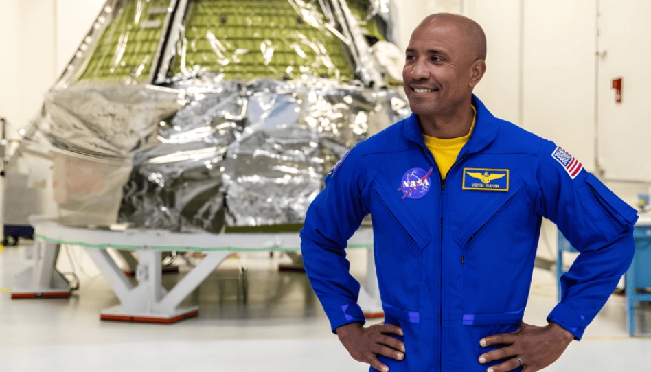 నాసా (NASA) బ్లూ స్పేస్‌సూట్ ధరించి, చిరునవ్వుతో స్పేస్‌క్రాఫ్ట్ (ఓరియన్ క్యాప్సూల్) ముందు నిలబడిన ఆర్టెమిస్ II మిషన్ పైలట్ విక్టర్ గ్లోవర్
